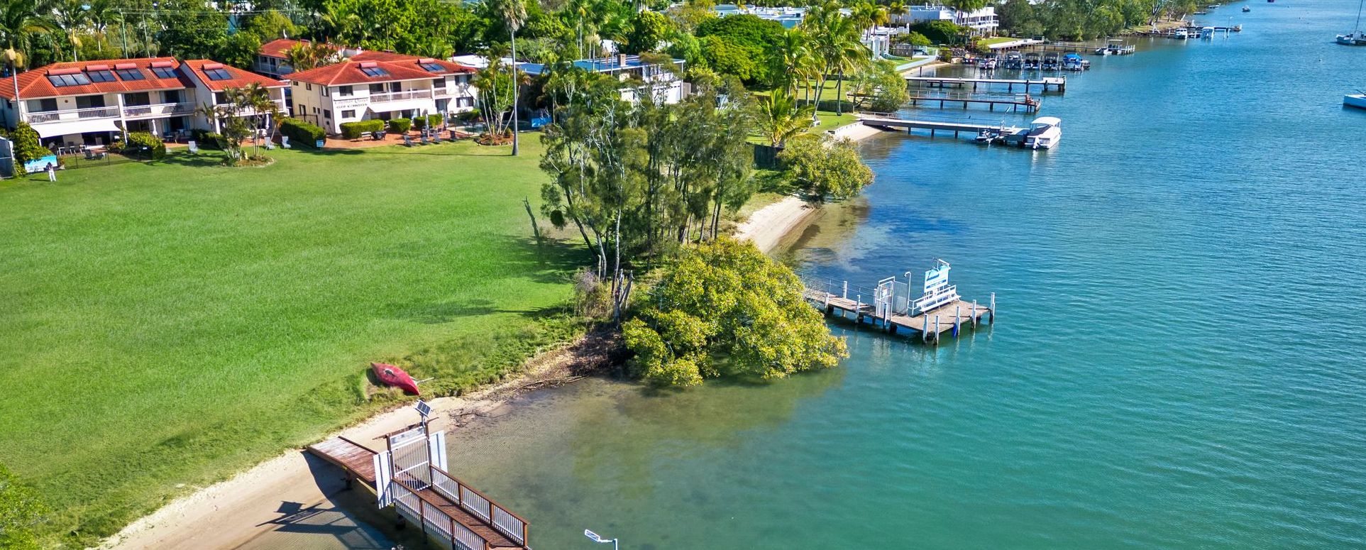 Private Guest Jetty and Waterfront Lawn High-angle aerial capture of Cayman Quays Noosa Riverfront resort highlighting the private guest jetty, expansive manicured grass lawn, and panoramic views of the water.