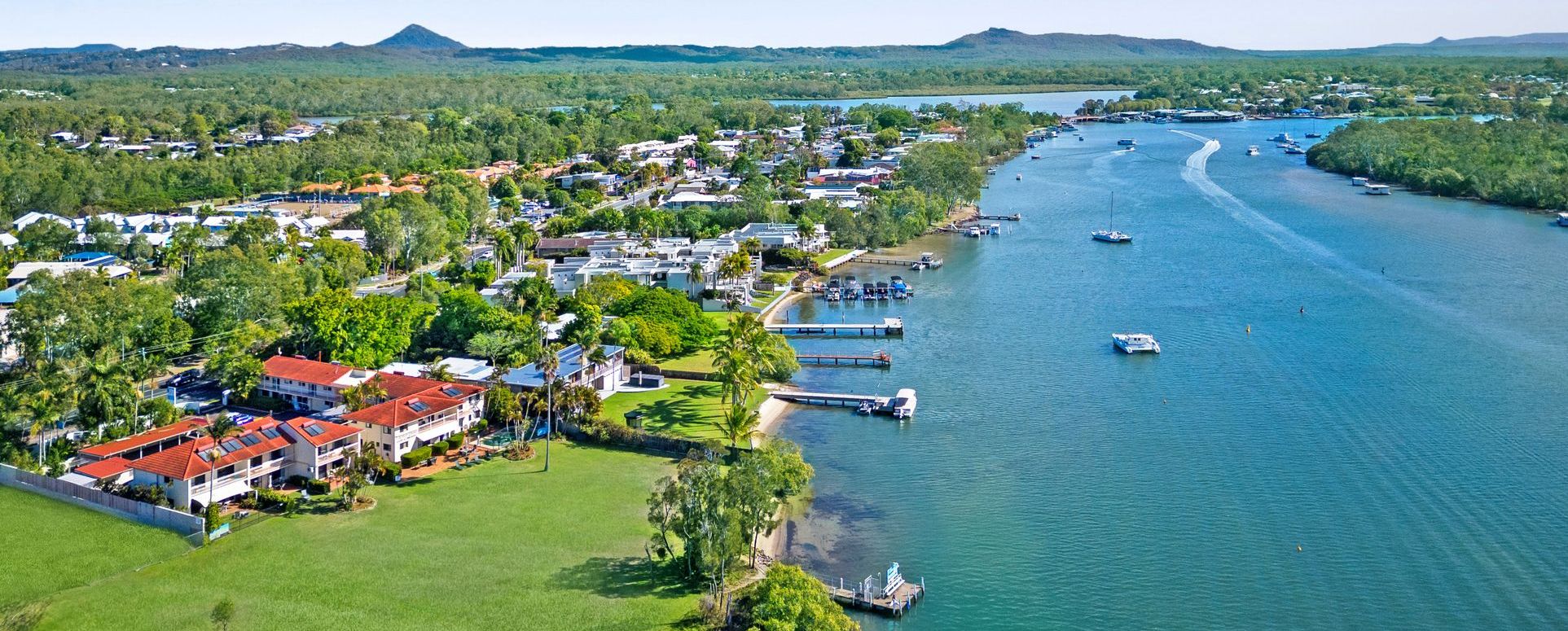 Cayman Quays Absolute Riverfront Aerial View Aerial view of Cayman Quays holiday apartments in Noosaville showing the 70m absolute riverfront grassland leading to the Noosa River, private guest boat mooring, and distant Sunshine Coast hinterland mountains.