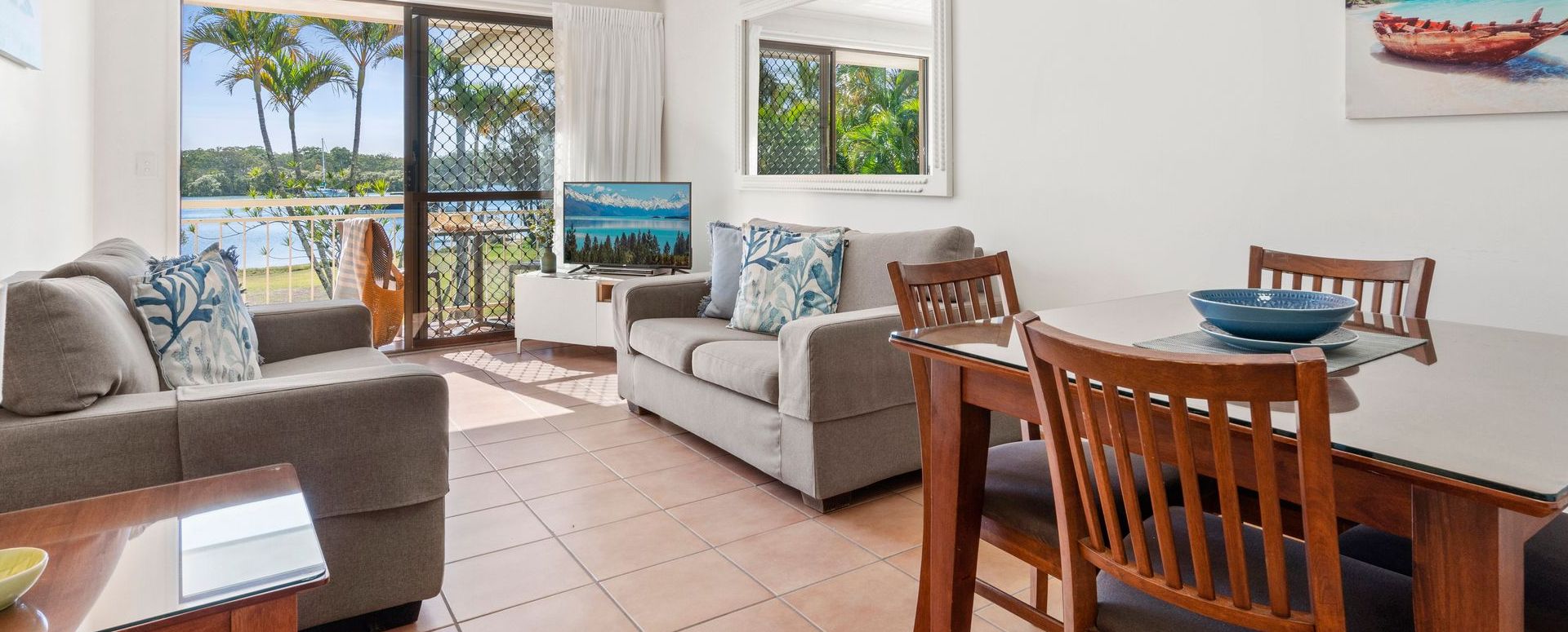 Apartment Living Dining Area and Patio Comfortably furnished apartment living room with a large flat-screen TV and dining table, looking out through glass sliders toward the private patio and the Noosa River.