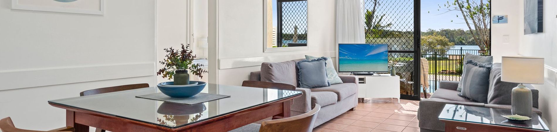 Riverside Apartment Open-Plan Living & Balcony View Wide open-plan view of the dining area and lounge room with a big screen TV, featuring glass sliding doors that open out to a private balcony with direct Noosa River views.