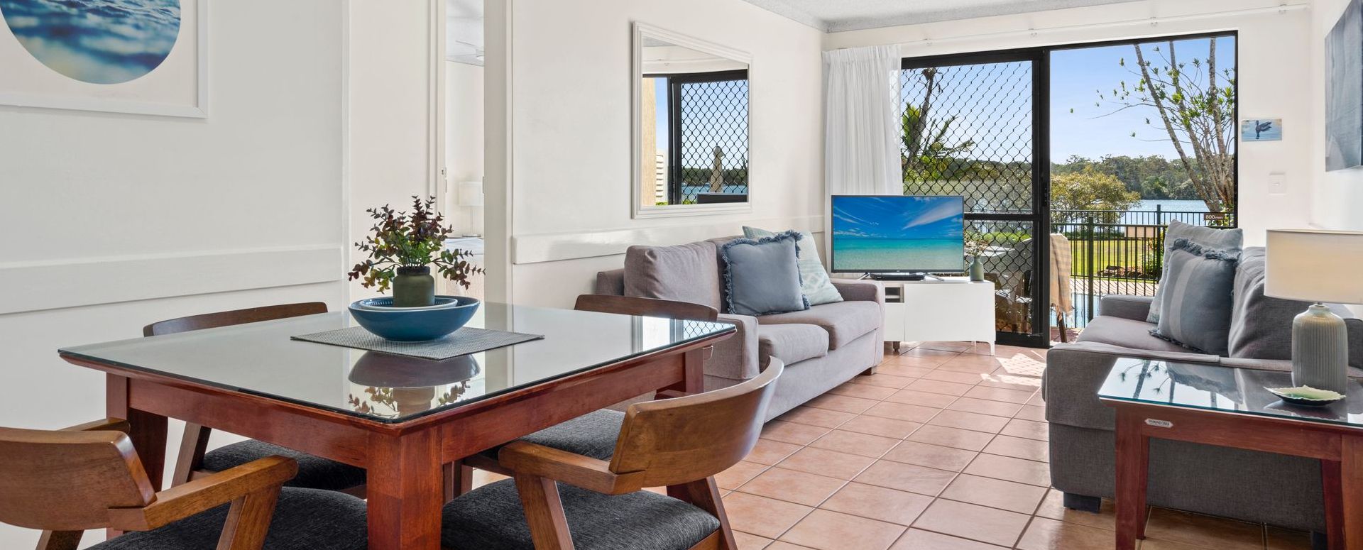 Self-Contained Apartment Living Area and Balcony View Fully self-contained holiday apartment interior at Cayman Quays featuring a modern lounge, big screen TV, dining area, and glass sliding doors opening to Noosa River balcony views.